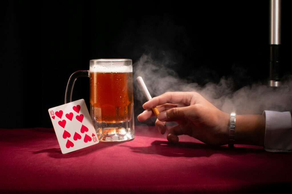 A hand holding a cigarette beside a beer and playing card on a red tablecloth.
